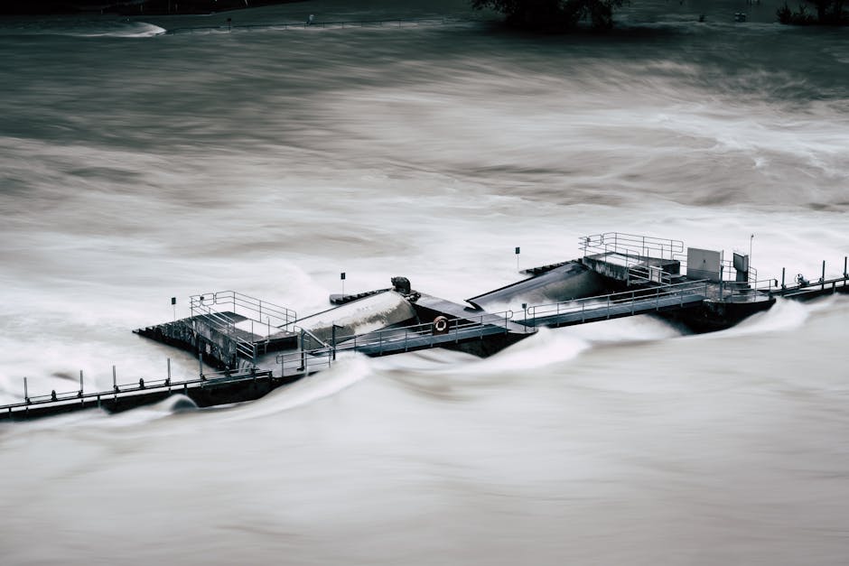 A residential street flooded with water covering the pavement and partially submerging a parked silver car, with trees and bushes around the area. In the background, there are multi-story apartment bu