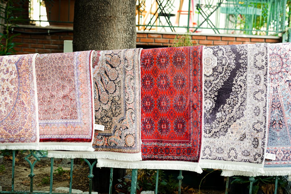 A collection of colorful, patterned rugs with intricate geometric designs and fringed edges hang on a dark wooden outdoor wall, illuminated by natural light. A wooden bench with a vibrant, floral-patt